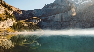 Mountain lake with clear reflection, surrounded by high, rocky mountains and pine trees, with light mist over the water.