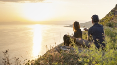 Zwei Personen sitzen bei Sonnenuntergang mit einem Glas Wein am Hang und blicken aufs Meer.