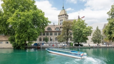 A long, narrow ferry on a river in front of a historic building and large trees.