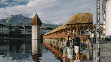 A pretty bridge with flowers, two people by the water, and mountains in the background.