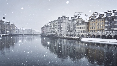 A snowy cityscape with historic buildings along the riverbank.