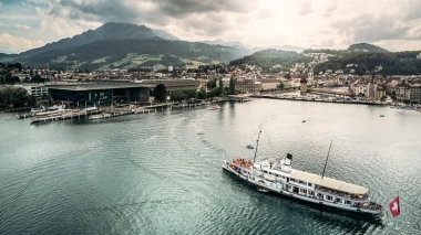 Eine Stadt am See mit Bergen im Hintergrund, ein Schiff fährt über das Wasser.