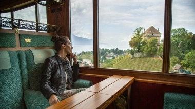 Eine Frau sitzt in einem Zugabteil und blickt nachdenklich aus dem Fenster auf eine Landschaft mit Weinbergen und einem Schloss.