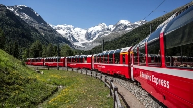 Ein roter Bernina Express Zug fährt durch eine alpine Landschaft mit schneebedeckten Bergen im Hintergrund.