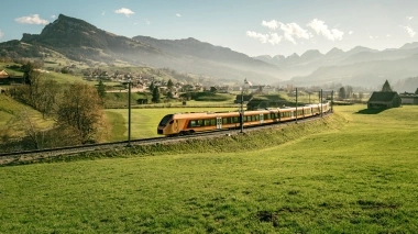 Ein Zug fährt durch grüne Hügel mit Bergen im Hintergrund.