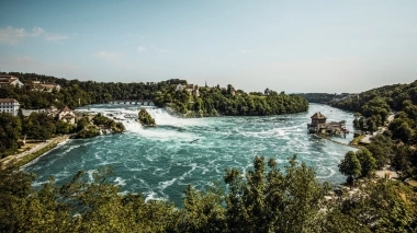 Eine weite Ansicht der beeindruckend rauschenden Rheinfälle inmitten grüner Landschaft unter klarem Himmel.