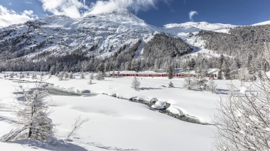 A red train travels through a snowy mountain landscape with a clear sky.