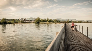 Eine Person schiebt ein Fahrrad über einen Holzsteg, mit Blick auf eine historische Stadt am See.