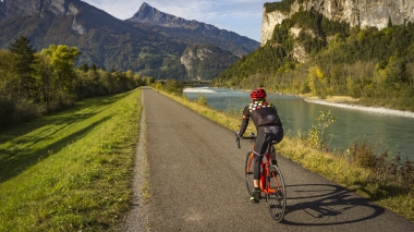 Ein Radfahrer auf einem Weg entlang eines Flusses mit Bergen im Hintergrund.