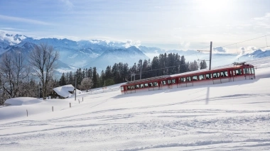 A red train travels through a snowy winter landscape with mountains in the background.