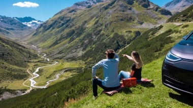 Two people sit on a meadow overlooking a valley in the mountains, next to a parked car.