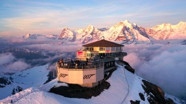 Eine Bergstation im Schnee mit 007-Logo, umgeben von schneebedeckten Alpen bei Sonnenuntergang.