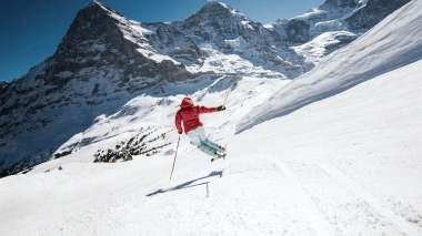 Uno sciatore in giacca rossa salta su una pista innevata con una straordinaria vista montana sullo sfondo.