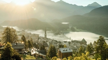 Sunset over an alpine town with a church, surrounded by mountains and a lake.