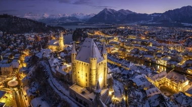 A winter city at night, illuminated buildings, snow-covered roofs, and mountains in the background.
