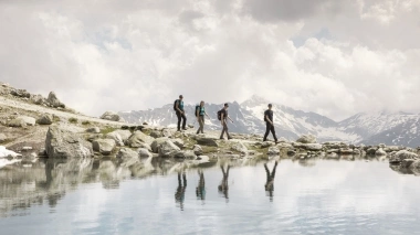 A group of hikers on a rocky path beside a mountain lake, with snow-capped mountains in the background.