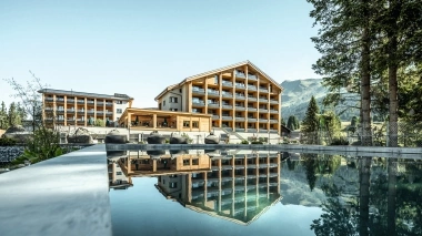 A modern hotel building is reflected in a calm pool, surrounded by mountains and trees.