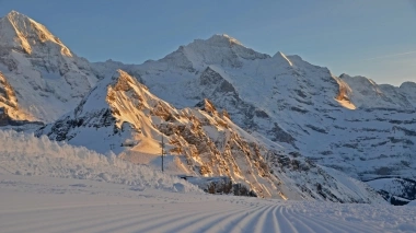 Sunlit, snowy mountain landscape with a groomed ski slope in the foreground.