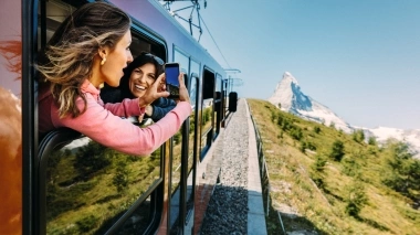 Two people leaning out of a train window taking a photo of a mountain landscape.