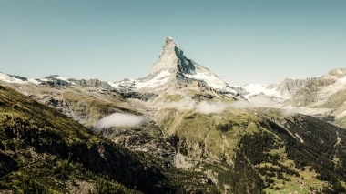 Eine Berglandschaft mit einer markanten Felsformation, umgeben von grünen Hügeln und Wäldern; im Hintergrund leicht bewölkter Himmel.