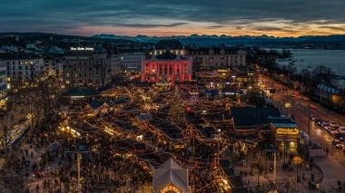 Aerial view of a festively lit Christmas market at dusk, surrounded by buildings with a view of mountains and a lake.