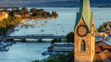 A church with a clock tower, view of the lake with boats and mountains in the background.