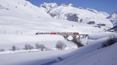 Un treno rosso attraversa un paesaggio invernale innevato, passando accanto a montagne innevate sotto un cielo limpido e azzurro.