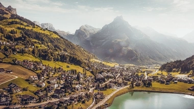 Villaggio con case vicino a un lago, circondato da colline verdi e alte montagne sullo sfondo.