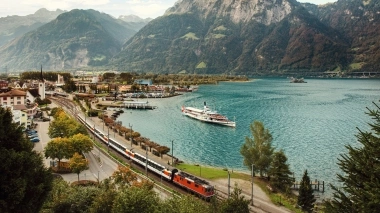 Paysage de montagne avec lac, train et bateau au premier plan.