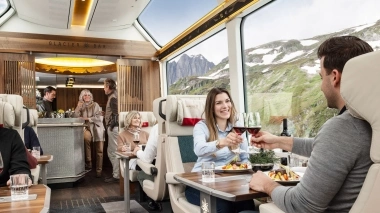 Innenansicht eines luxuriösen Panoramazugs mit grossen Fenstern; Reisende geniessen ein elegantes Abendessen mit Blick auf die alpine Landschaft.