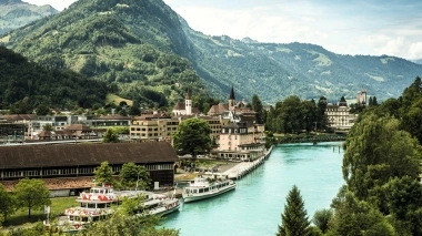 Vue de la ville avec rivière, bateaux et montagnes environnantes en arrière-plan.