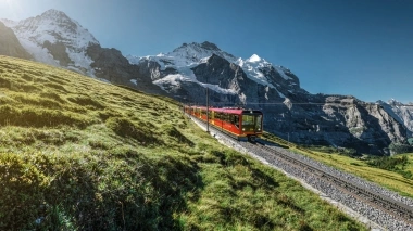 Eine rote Bahn fährt durch eine grüne Berglandschaft mit schneebedeckten Gipfeln im Hintergrund.