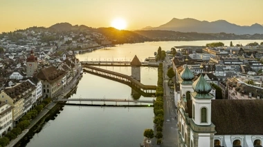 Sunset over a city with a bridge and river, mountains in the background.