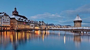 A historic bridge and buildings by the river at dusk, covered with snow.