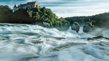 Wasserfall mit Schloss im Hintergrund, umgeben von üppiger Vegetation.