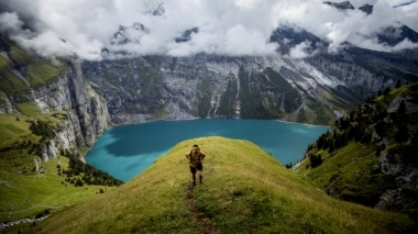 A person hikes on a green hill with a turquoise mountain lake in the background.