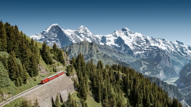 Eine rote Bahn fährt durch grüne Berge mit schneebedeckten Gipfeln im Hintergrund.