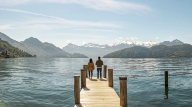 Two people stand on a wooden pier looking over a lake at snow-covered mountains.