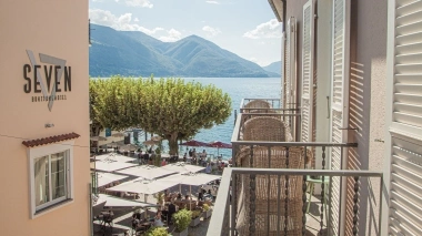 Blick von einem Balkon auf einen Platz mit Tischen und Stühlen am See, umgeben von Bergen.