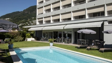 Hotel building with balconies, pool in the foreground, and green hills in the background.