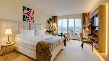 An elegant hotel room with a double bed, artwork on the wall, and a large window with a mountain view.