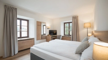 A bright hotel room with a double bed, wooden desk, chair, TV, and large windows with curtains.