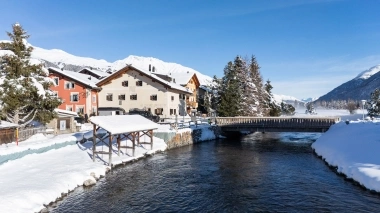 A winter mountain landscape with houses by the river and snow-covered mountains in the background.