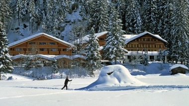 A person on skis in a snowy field in front of a large wooden building in a snow-covered forest.