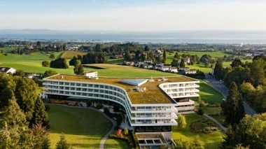 Modern building with a green roof in a rural setting, overlooking a lake in the distance.