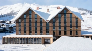 Un bâtiment moderne à plusieurs étages avec une façade en bois et un toit enneigé dans un paysage alpin.