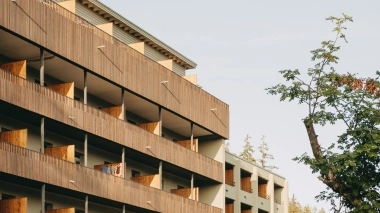 Multi-story building with wooden balconies, next to a tree against a blue sky.