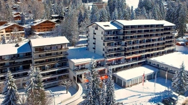 Luftaufnahme eines grossen Gebäudekomplexes in einer schneebedeckten Landschaft mit Tannenbäumen.