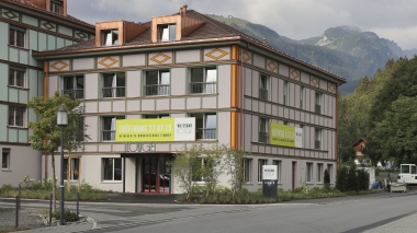 Façade d'un immeuble à plusieurs étages avec une banderole d'ouverture et des montagnes en arrière-plan.