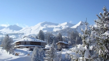 A winter mountain landscape with heavily snow-covered trees and houses in the foreground.